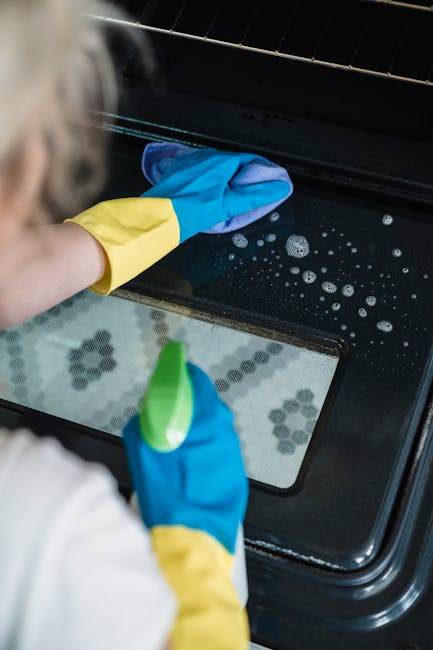 A person wearing blue and yellow rubber gloves is deep cleaning the interior of a black oven, using a cloth to wipe the oven door and a spray bottle in hand. The oven's glass door reflects the light,