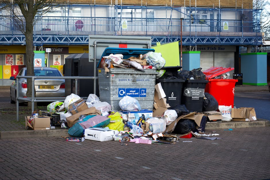 A patch of ground behind a black metal fence is littered with various types of waste, including crumpled paper, plastic bags, and food packaging. The waste appears scattered and mixed with natural deb